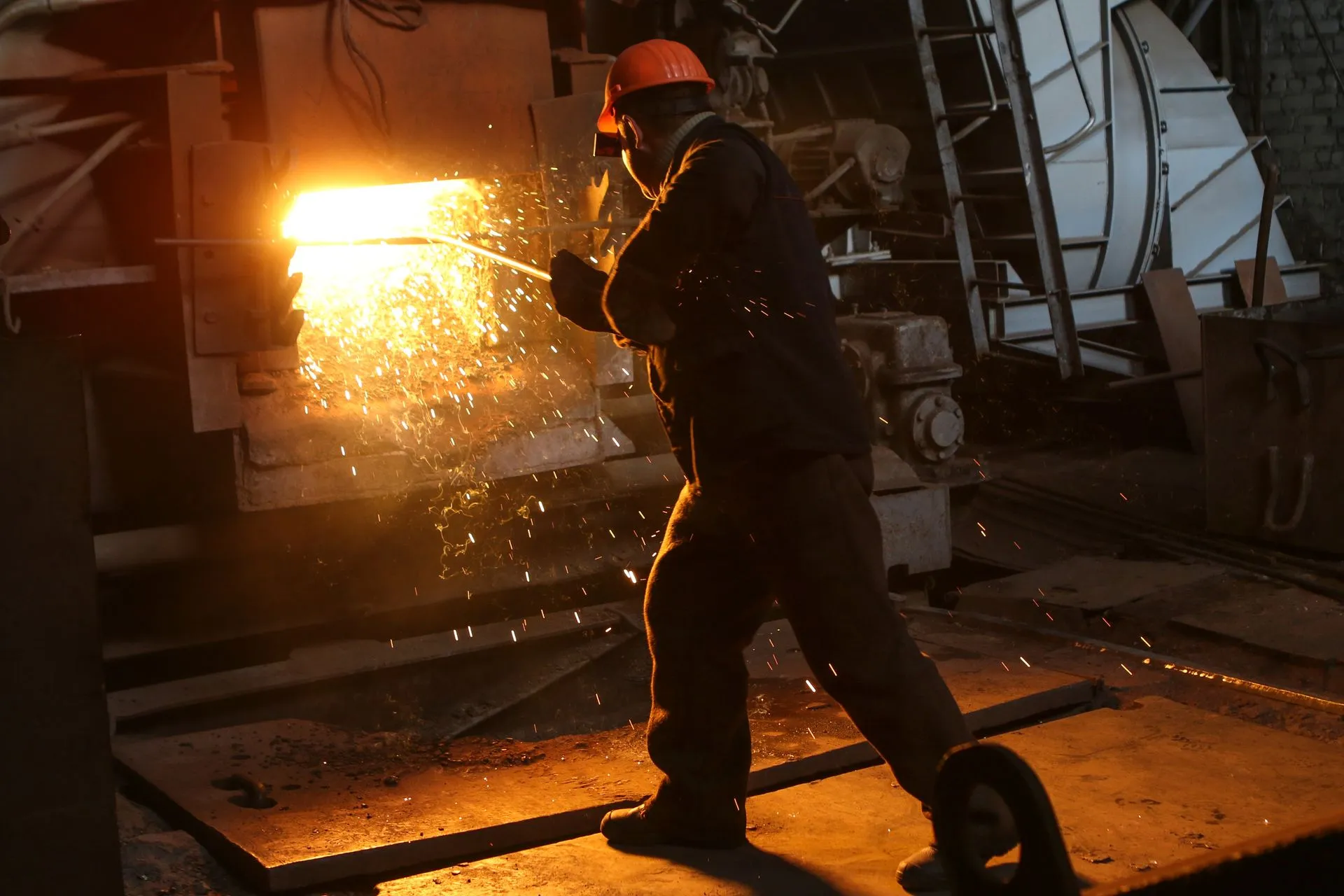 Trabajador con casco naranja atendiendo un horno de fundición entre chispas